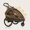 A brown and gray child trailer for cycling or walking, shown from a side-diagonal angle against a light beige background. Orange accents highlight the trailer's frame and seams. Arrows indicate a curved motion or airflow. Produktbild-Vorschau
