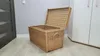 A wicker chest with its lid open, placed on a light wooden floor next to a gray wall and a white cabinet. Voir les images du produit