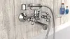 A chrome bathtub faucet with a handheld shower attachment is mounted on a light wood-paneled wall above a white bathtub. Two blue bottles of toiletries are on a small white shelf to the right. Náhled obrázku produktu
