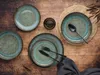 A rustic table setting with plates, cutlery, and a small glass of liquid. Náhľad obrázku produktu
