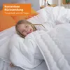 A young girl is lying in a bed covered by a white comforter, with orange pillows in the background. Produktbild-Vorschau