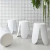 Four white stools with cream-colored seats are arranged on a light gray floor in front of a white grid-patterned wall. Green plants hang from the top right. Produktbild-Vorschau