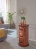 A round wooden side table with three drawers is placed in a living room setting, next to a sofa and a large window with sheer curtains. Decorative items, including a gold vase with greenery and a gold acorn-shaped object, are placed on top of the table. A  Podgląd zdjęcia produktu