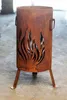 A rusty metal fire pit with flame cutouts and three legs, placed on a concrete floor. Produktbild-Vorschau
