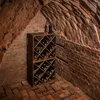 A dark, rustic wine cellar with brick walls and floor, featuring a wooden wine rack filled with bottles. Voir les images du produit