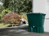 A woman sits on a bench reading a book in a garden next to a green rain barrel. Voir les images du produit