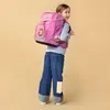 A young girl wearing a pink backpack and jeans, posing against a plain background. Voir les images du produit