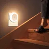 A person's foot in a black slipper is stepping onto a wooden stair, illuminated by a wall-mounted LED night light. Podgląd zdjęcia produktu
