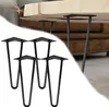 Four black metal hairpin legs are arranged in the foreground, with a wooden tabletop and sofa visible in the background. Produktbild-Vorschau