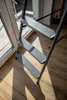 A close-up view of a metal step stool with three steps, placed on a wooden floor near a window with sunlight casting shadows. Voir les images du produit