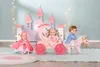 Three dolls are arranged in a playful scene with a pink castle backdrop, a pink carriage, and a pink horse. One doll is seated in the carriage, another is riding the horse, and a third is standing to the side wearing a pink cape. Produktbild-Vorschau