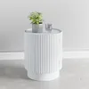 A white round side table with a fluted design, a potted plant, and a glass of water on top, set against a light gray wall and a concrete floor. Anteprima dell’immagine prodotto