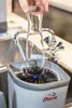 A hand is holding a glass carafe over a brush-like cleaning device in a sink. Podgląd zdjęcia produktu
