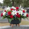 A floral arrangement in a black pot with wooden legs sits on a gravestone in a cemetery. Podgląd zdjęcia produktu