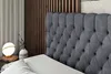 A close-up view of a dark gray upholstered headboard of a bed, with a white pillow in the foreground. A bedside table with a lamp and a vase is visible to the left, and a slatted wooden wall is in the background. Produktbild-Vorschau
