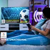 A couple is sitting on the floor in front of a TV, watching a soccer game. The TV screen displays a soccer game with DAZN branding. There are also overlays indicating Google Home compatibility and voice control. Produktbild-Vorschau