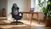 A black gaming chair is placed next to a wooden desk with a laptop on it, in a well-lit room with plants. Anteprima dell’immagine prodotto
