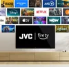 A wall-mounted television displaying various streaming service logos and the JVC and Fire TV logos, with a wooden media console below. Produktbild-Vorschau