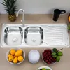 A stainless steel double sink with a faucet, a pineapple, and bowls of fruit on a countertop. Produktbild-Vorschau
