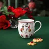 A Christmas-themed mug with a snowman design sits on a green tablecloth, next to two festive cookies. Red flowers and berries are visible in the background, along with a blurred Christmas tree. Voir les images du produit
