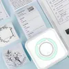 Weißer tragbarer Thermodrucker mit hellgrünem Akzent, der ein Versandetikett und dekorative Bilder auf einer hellblauen Oberfläche druckt Náhled obrázku produktu