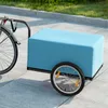 A blue bicycle cargo trailer is attached to a bicycle, parked on a paved surface next to a grassy area. Produktbild-Vorschau