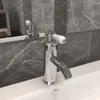 A close-up shot of a chrome-plated bathroom faucet with a white handle, set against a marble-tiled wall and a white sink. Podgląd zdjęcia produktu