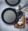 Two frying pans are shown on a light gray surface, with a wooden cutting board, onions, garlic, and green onions in the background. Náhled obrázku produktu
