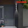 A modern dark gray front door with a long silver handle and a potted lavender plant next to it. A textured dark gray wall with a white LED wall light is to the right of the door. Green foliage is visible at the top right. Produktbild-Vorschau