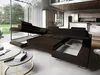 A modern living room with a large sectional sofa that has its storage compartments open, revealing the bed mechanism. Large windows overlook a garden, and a kitchen area is visible in the background. Produktbild-Vorschau