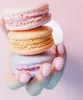 A hand with pink nail polish is holding a stack of three macarons in pastel colors: pink, peach, and lavender. Anteprima dell’immagine prodotto