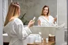 A woman in a bathrobe is holding a bottle of skincare product in front of a mirror in a bathroom. Produktbild-Vorschau