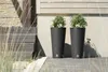 Two tall, dark grey planters with green plants sit on a wooden deck next to a light-colored wooden wall. Produktbild-Vorschau