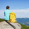 A person is sitting on a rock in the mountains with a backpack and trekking poles. Produktbild-Vorschau
