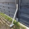 A metal storm anchor is attached to a dark wooden fence post and anchored to the ground. Produktbild-Vorschau