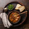 A rustic wooden table setting with a bowl of hearty soup, bread, and herbs. Produktbild-Vorschau