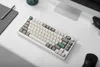 A white mechanical keyboard is placed on a white desk next to a pen and a notebook. Produktbild-Vorschau