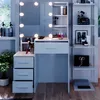 A white vanity table with LED lights and drawers, set up in a room with a window and shelves. Náhled obrázku produktu