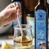 A hand is stirring a clear liquid in a glass jar with a wooden spoon. A bottle of oil is visible on the right side of the image, and a slice of lemon is in the foreground. Produktbild-Vorschau