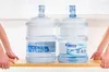 Two large water bottles on a wooden surface, held by two people from the sides. Produktbild-Vorschau