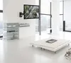 A modern living room with a TV mounted on the wall, a glass shelf unit, and a low platform bed with a rug. Podgląd zdjęcia produktu