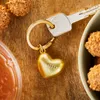 A close-up shot of a golden heart-shaped keychain with a key, placed on a wooden surface, with some chocolates and a jar of jam visible. Produktbild-Vorschau