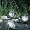 Three solar-powered rock lights are placed among rocks and tall green plants in a garden setting. Produktbild-Vorschau