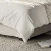 A close-up view of a bed with white bedding and a textured rug on the floor. Produktbild-Vorschau