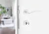 A close-up view of a white door with a white door handle and a key in the lock. The background is blurred and shows a living room. Produktbild-Vorschau