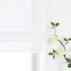 A close-up view of a white Roman blind with a branch of artificial flowers with light yellow blossoms and green leaves placed to the right against a white wall. Produktbild-Vorschau