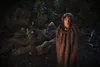 A group of people wrapped in blankets in a dark forest setting, with one person standing in the foreground looking at the camera. Podgląd zdjęcia produktu