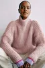 A woman wearing a pink knitted sweater and white pants, sitting cross-legged. Produktbild-Vorschau