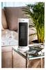 A white tower heater is placed on a glass table next to a sofa and a plant. Podgląd zdjęcia produktu