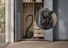 A vacuum cleaner is stored inside a closet with drawers and hanging space. A close-up inset shows the vacuum's attachments. Produktbild-Vorschau